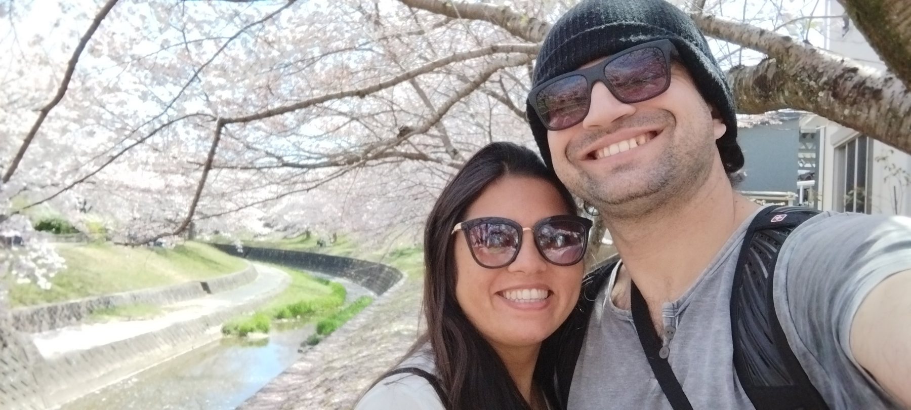 Silvana e o marido sorrindo sob cerejeiras em flor, ao lado de um rio